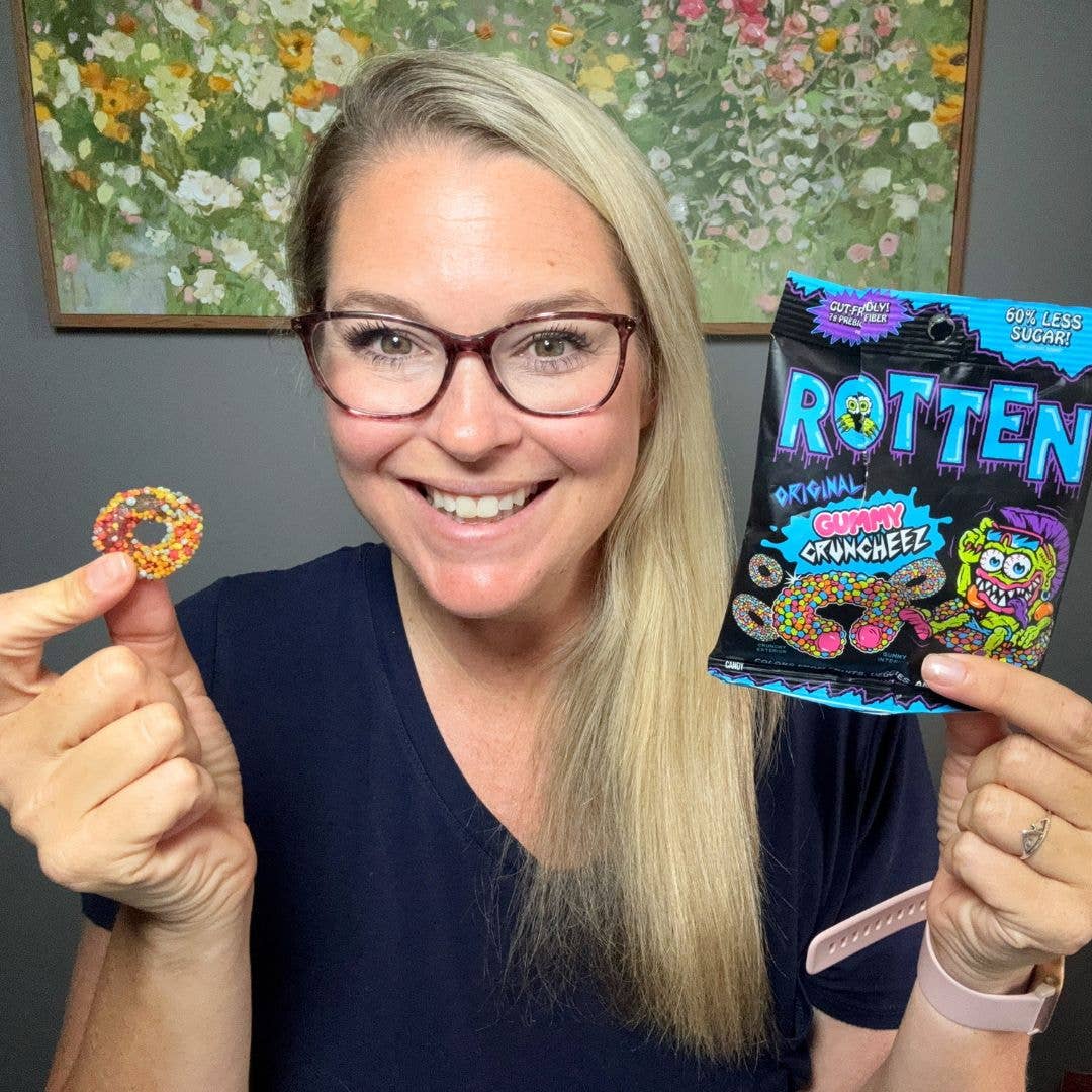 Woman holding a colorful donut and a 'Rotten' candy package in front of a floral painting.